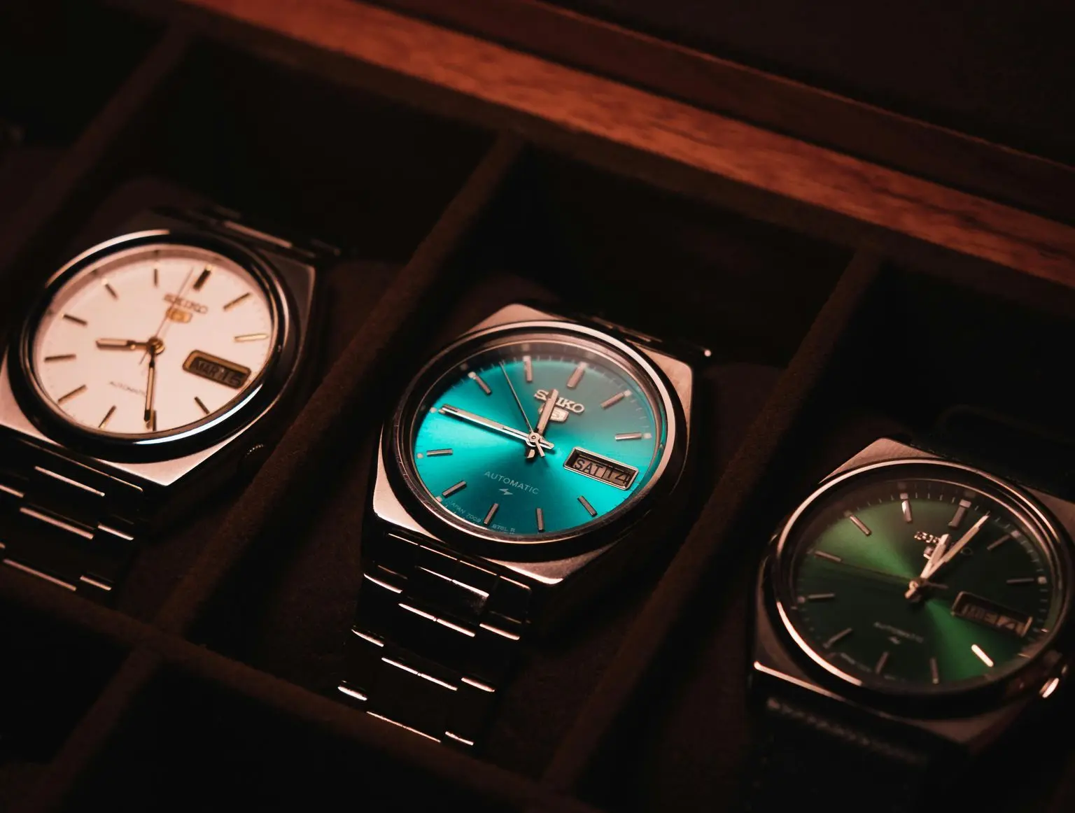 Three wristwatches with different colored faces—white, blue, and green—are displayed side by side in a dark, padded watch case.