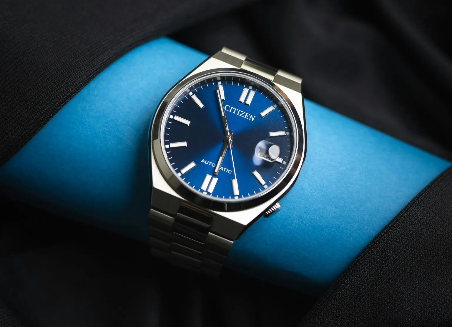 A Citizen analog wristwatch with a blue face, white hour markers, a date window, and a metal band displayed on a blue cylindrical surface.