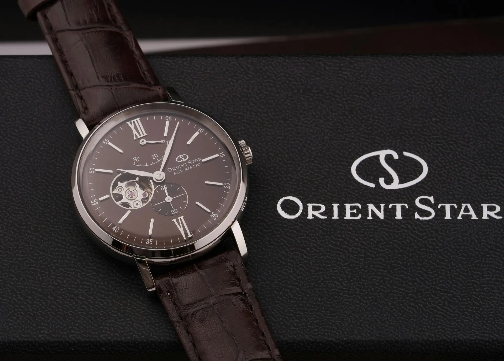 A brown leather strap Orient Star wristwatch with an open heart dial rests on a black Orient Star box featuring the brand’s logo and name.