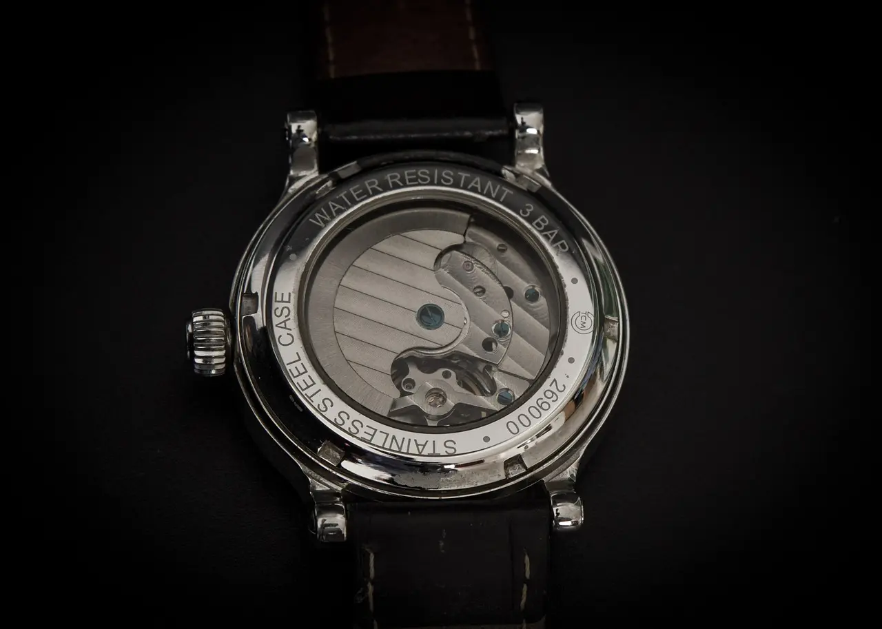 Close-up of a wristwatch case back showing its mechanical movement, labeled "Water Resistant 3 Bar" and "Stainless Steel Case" with a leather strap.