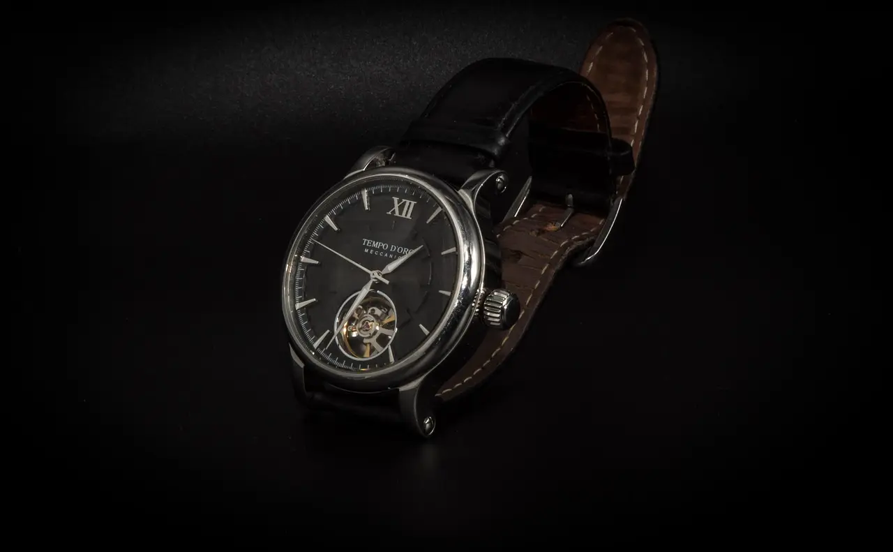 A black leather wristwatch with a silver case, black dial, Roman numerals, and an exposed mechanical movement is displayed on a dark background.