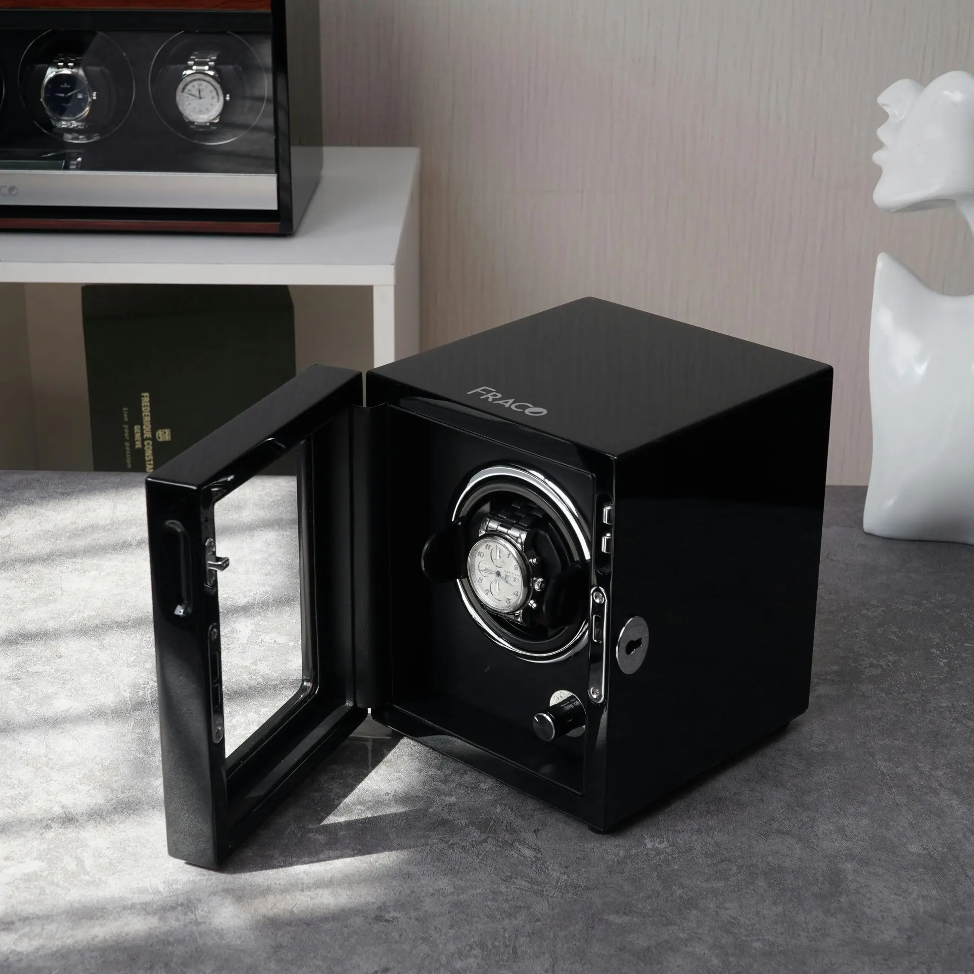lack Watch Winder Safe A black watch winder safe with a glass door is open on a gray surface, displaying a wristwatch inside. A watch case and abstract white sculpture are in the background.
