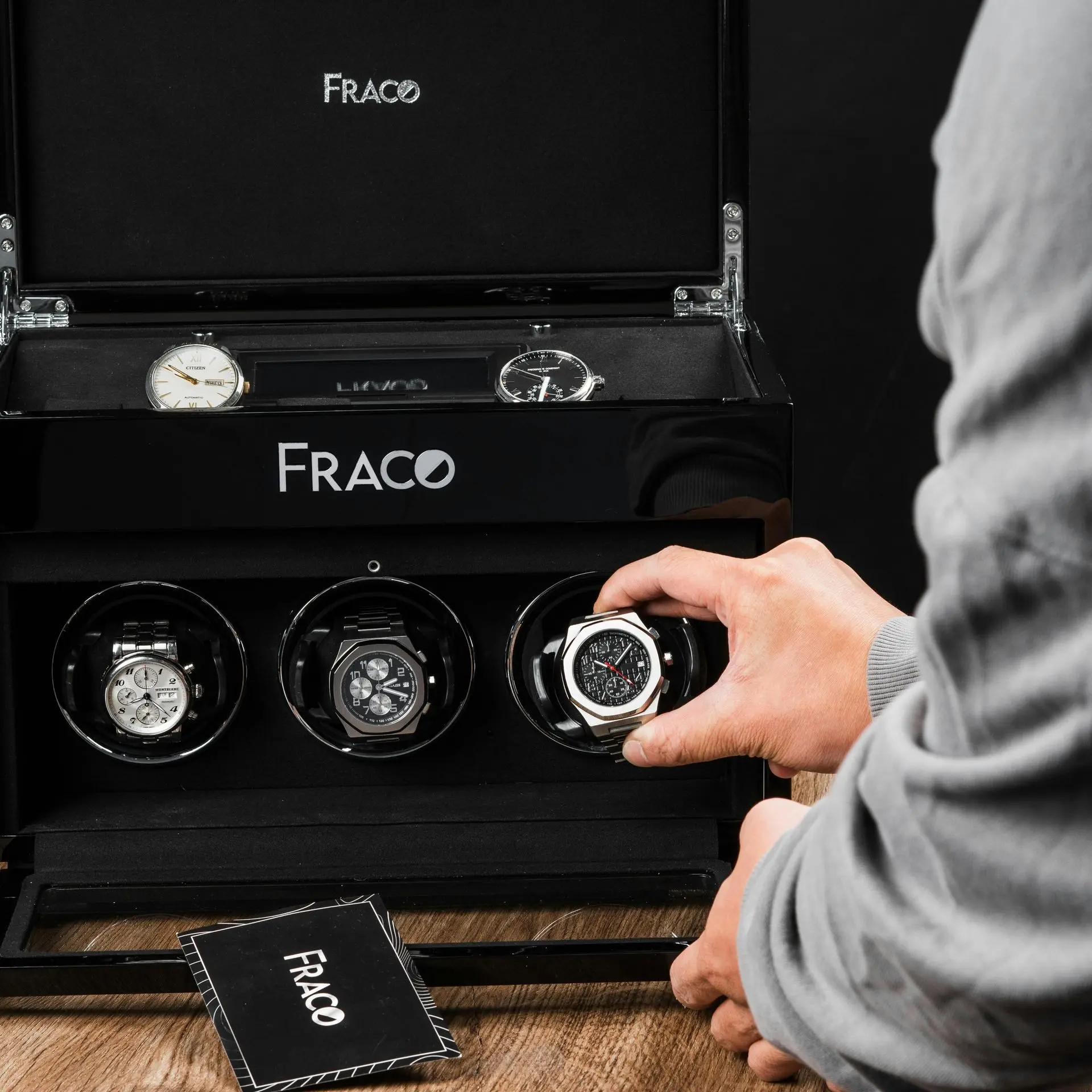 Black Watch Winder Cabinet with Watches A person places a wristwatch into a black FRACO watch winder case, which contains multiple watches and an instruction booklet on a wooden surface.