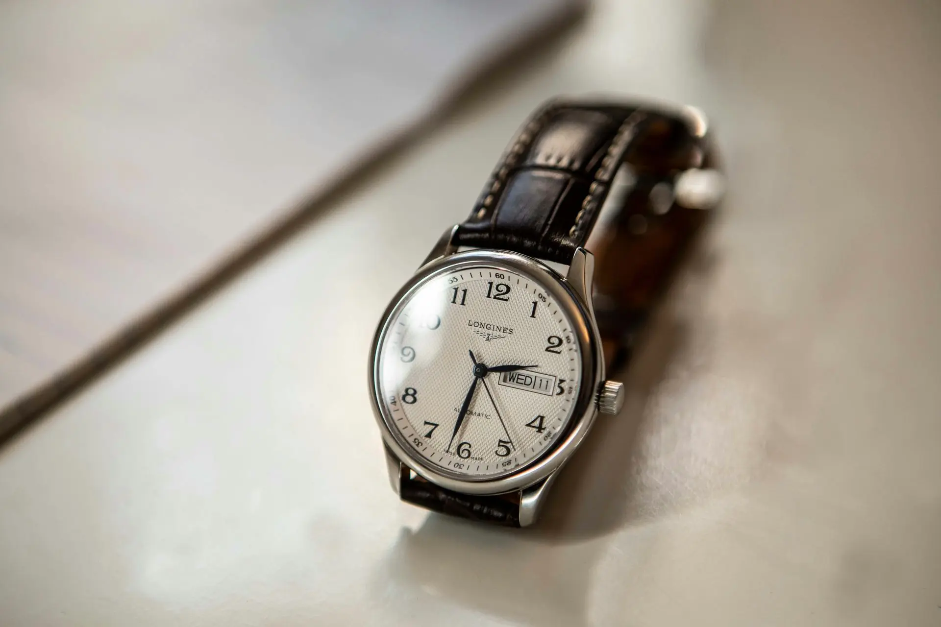 A classic Longines wristwatch with a white dial, black Arabic numerals, and a brown leather strap resting on a light surface