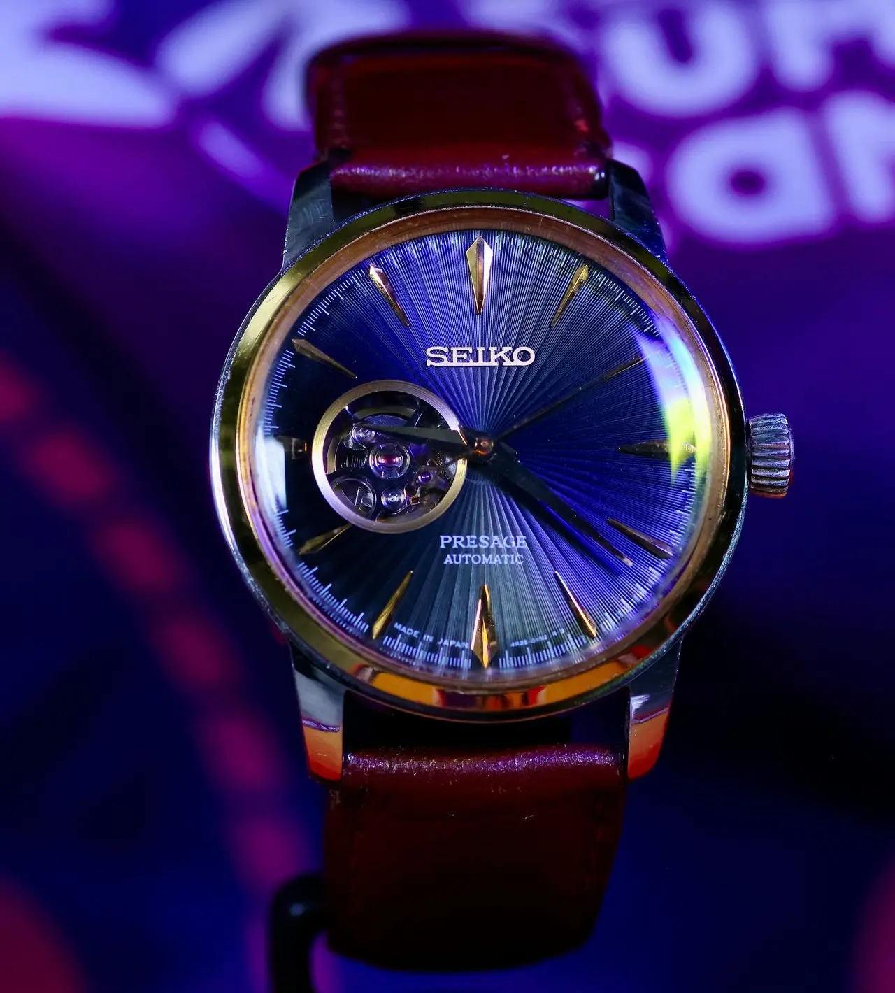 Close-up of a Seiko Presage automatic watch with a blue sunburst dial, visible open-heart aperture showing a Japanese movement, gold-toned indices, and a brown leather strap.