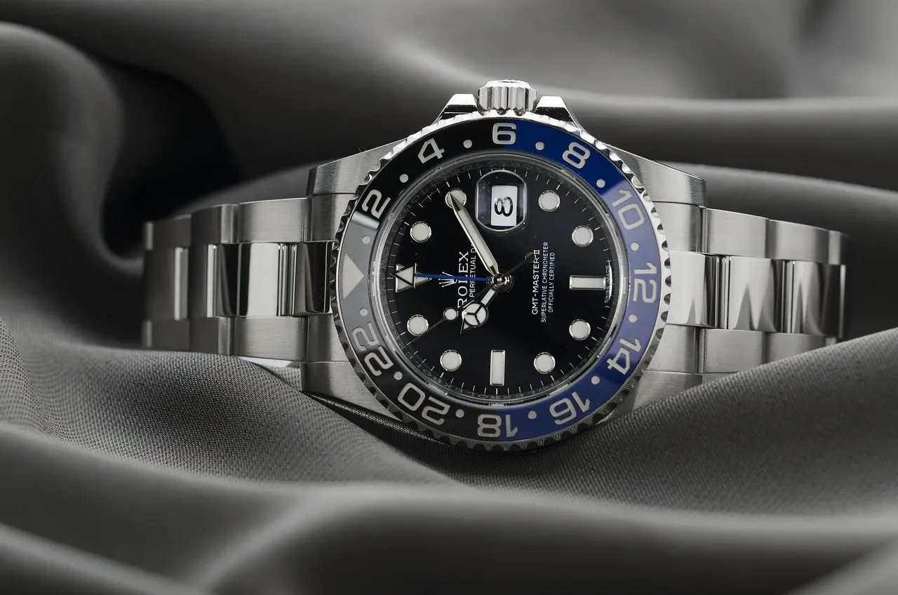 A silver Rolex GMT-Master II watch with a black and blue bezel and metal bracelet is resting on a gray satin fabric.