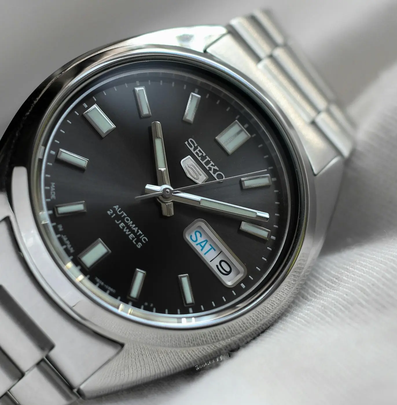 A silver Seiko 5 automatic wristwatch with a black face, displaying the day and date as Saturday the 9th, resting on a light-colored fabric.