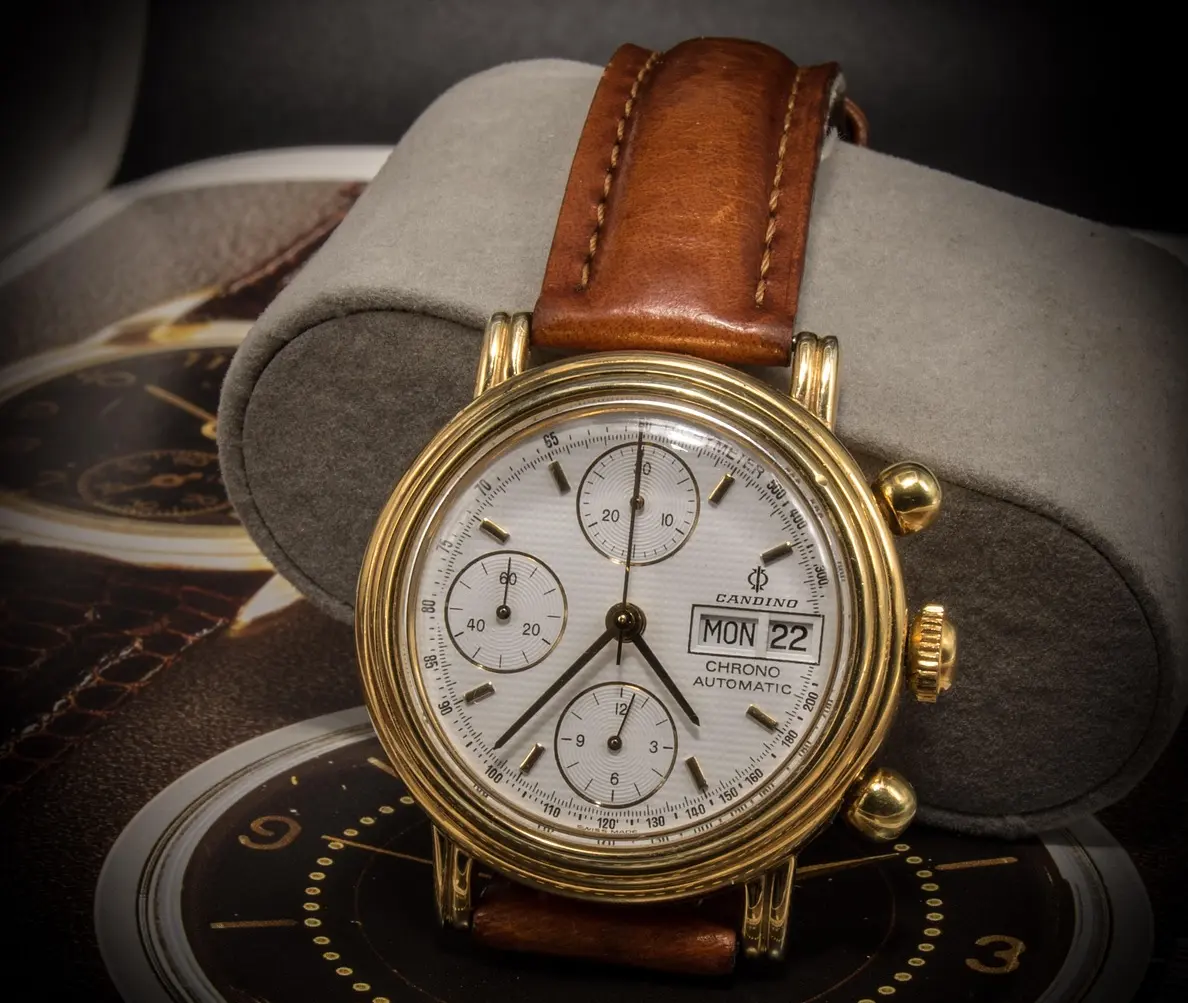 Gold-tone chronograph wristwatch with a white dial, multiple subdials, day/date window, and brown leather strap.