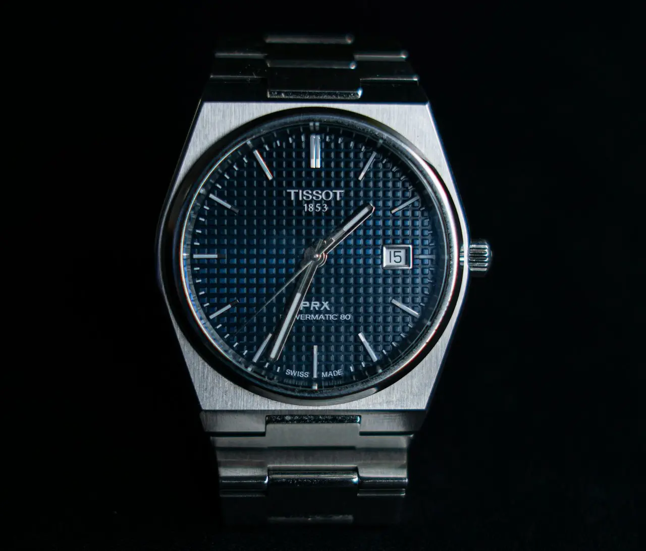 Tissot PRX Powermatic 80 A silver Tissot wristwatch with a blue textured dial, silver hour markers, metal strap, and a date display set to the 15th, photographed against a black background.