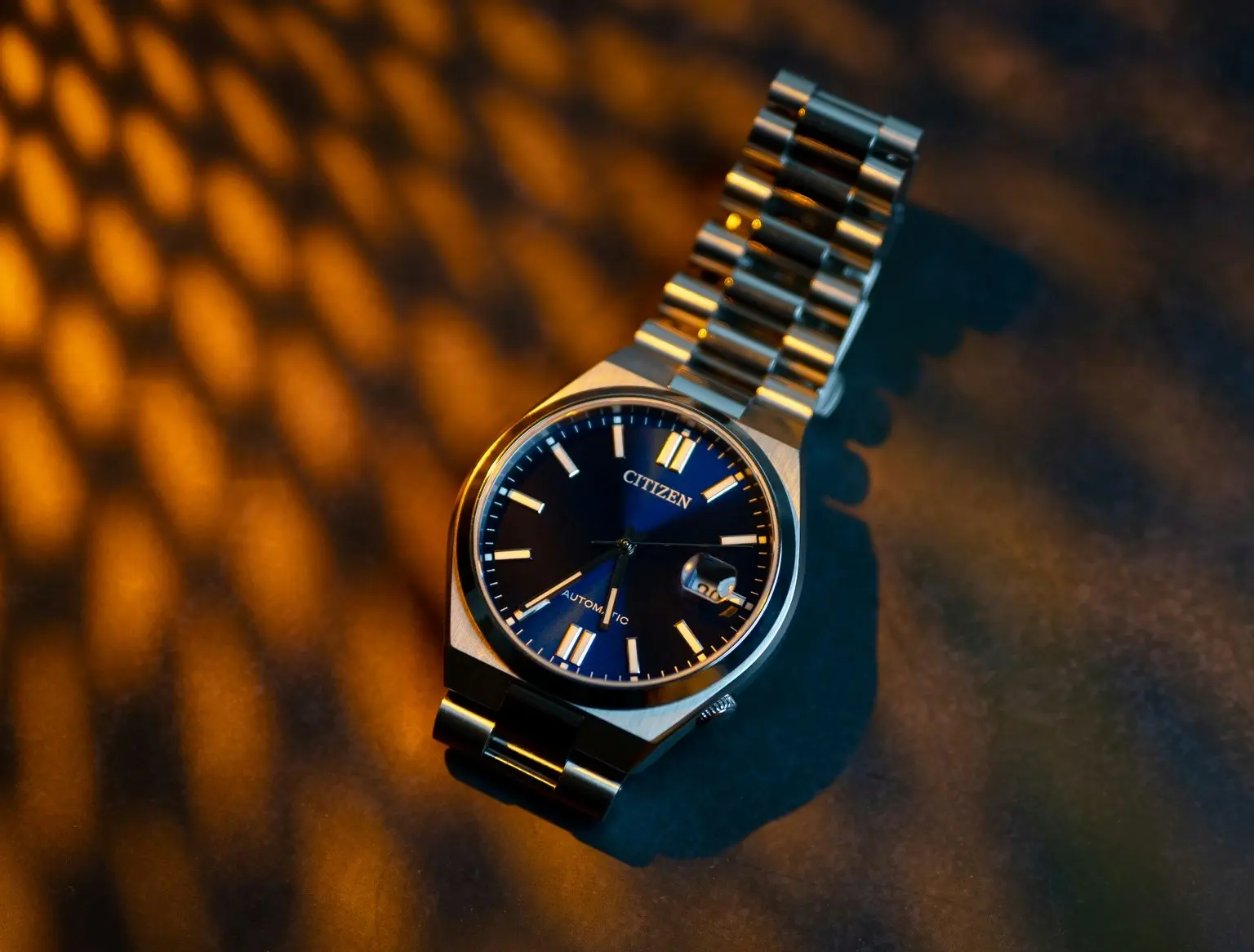 Citizen Tsuyosa A Citizen wristwatch with a silver metal band and a black dial is placed on a surface with warm, patterned lighting.