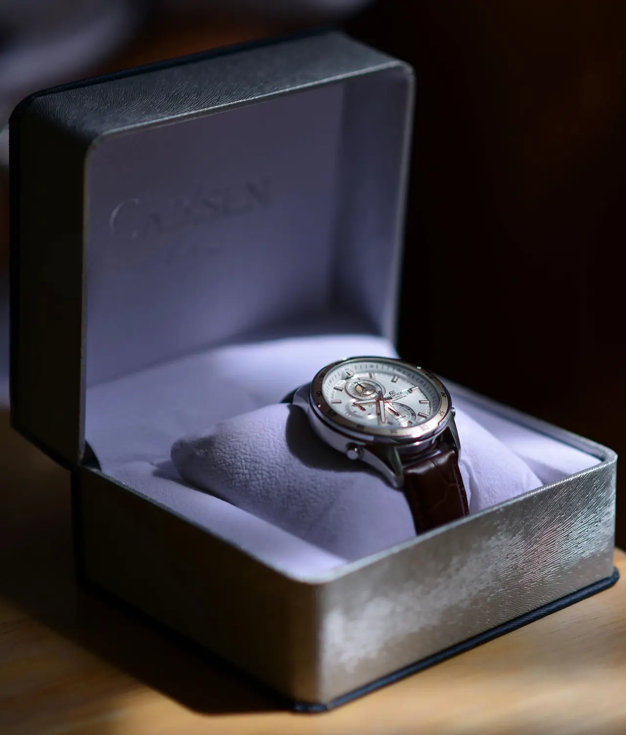 A wristwatch with a brown leather strap and silver casing is displayed in an open cushioned box with a soft interior.