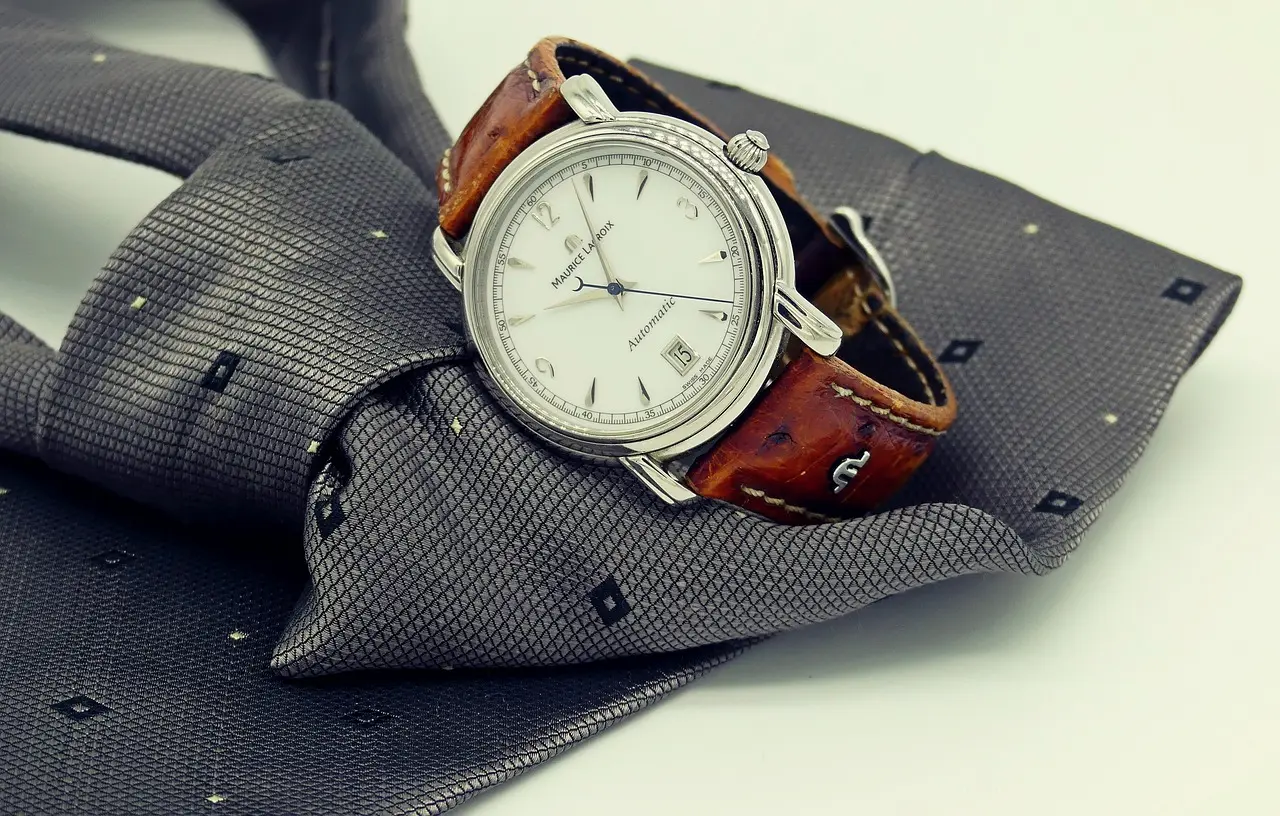 A wristwatch with a brown leather strap rests on a folded gray patterned necktie, both placed on a white surface.