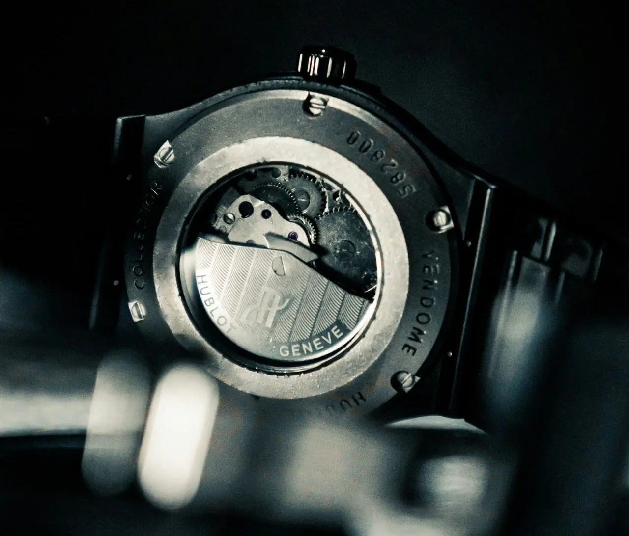 Dark, moody close-up photograph of the caseback of a Hublot wristwatch, showing the exhibition case window displaying the automatic movement's rotor and internal gears.