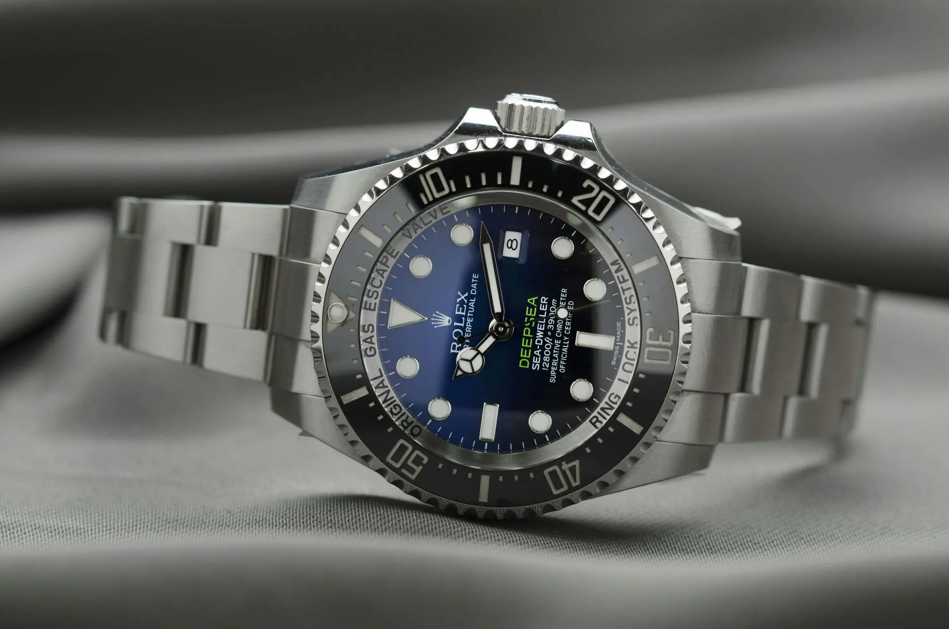 Close-up of a stainless steel Rolex Deepsea diver's watch with a blue dial and black bezel, resting on a gray fabric surface.