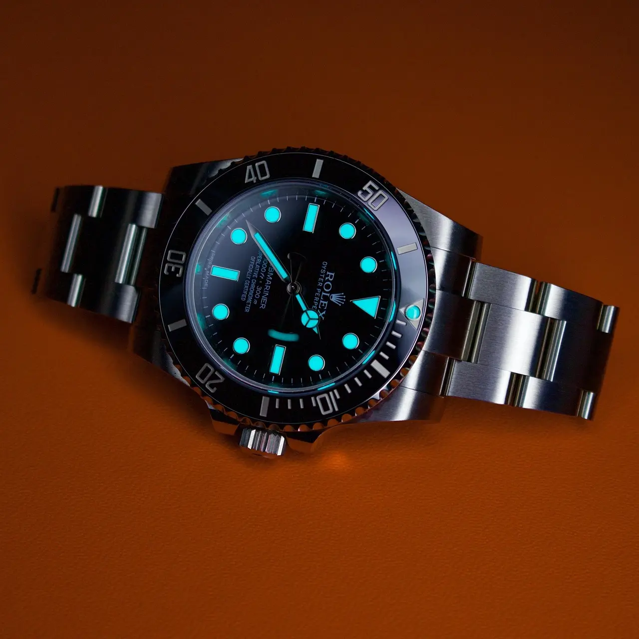 Luminescent diver's watch with steel bracelet on orange backdrop Close-up of a Rolex-style diver's watch with a black dial, glowing blue lume on the hands and markers, and a metal bracelet on an orange background.