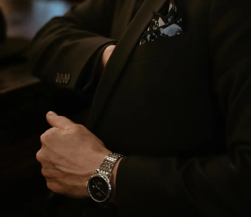 A person in a dark suit with a patterned pocket square adjusts their sleeve, wearing a silver wristwatch.