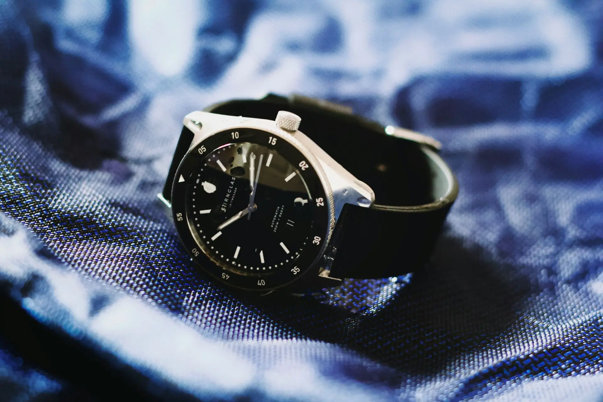 Minimalist watch with clean black dial and slim silver case resting on blue fabric, showcasing modern simplicity