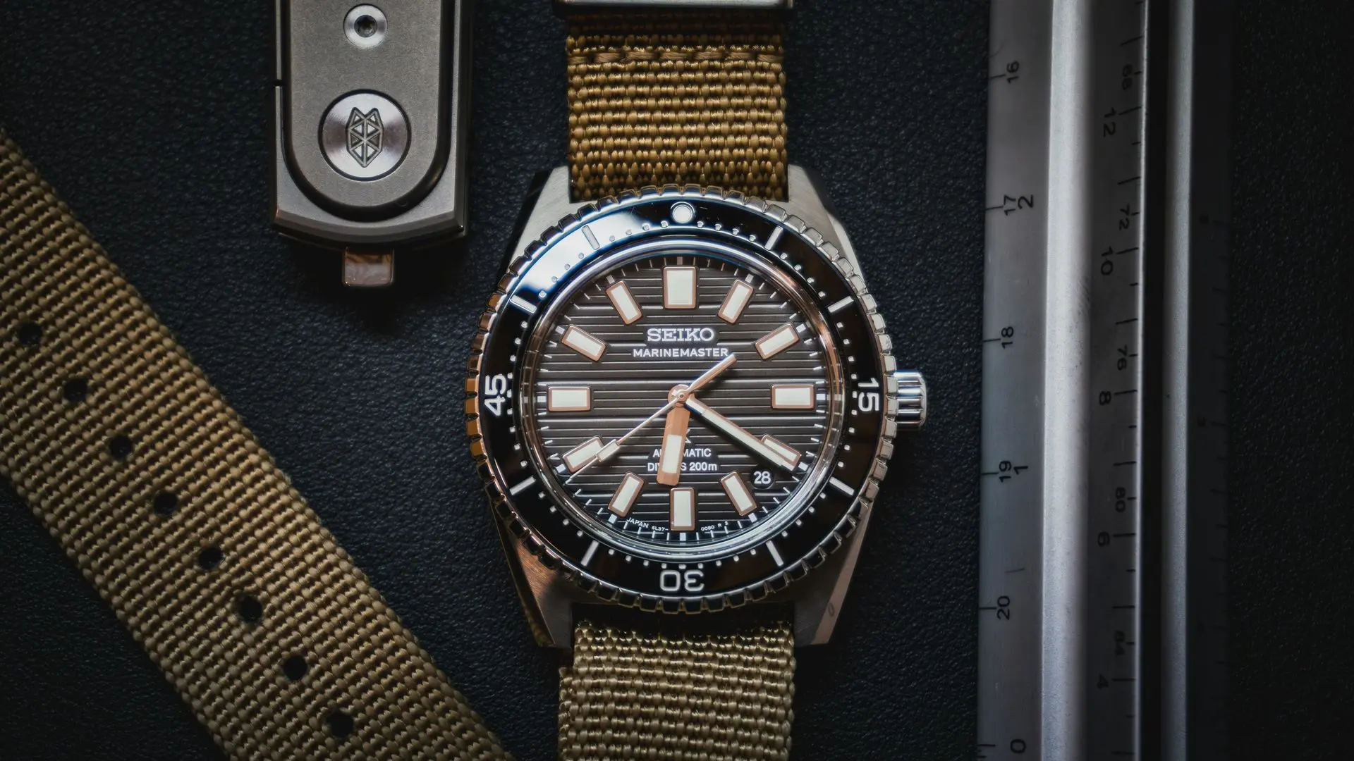 Seiko diver’s watch with a rugged tan fabric strap resting beside a metal ruler and case, highlighting water resistance.