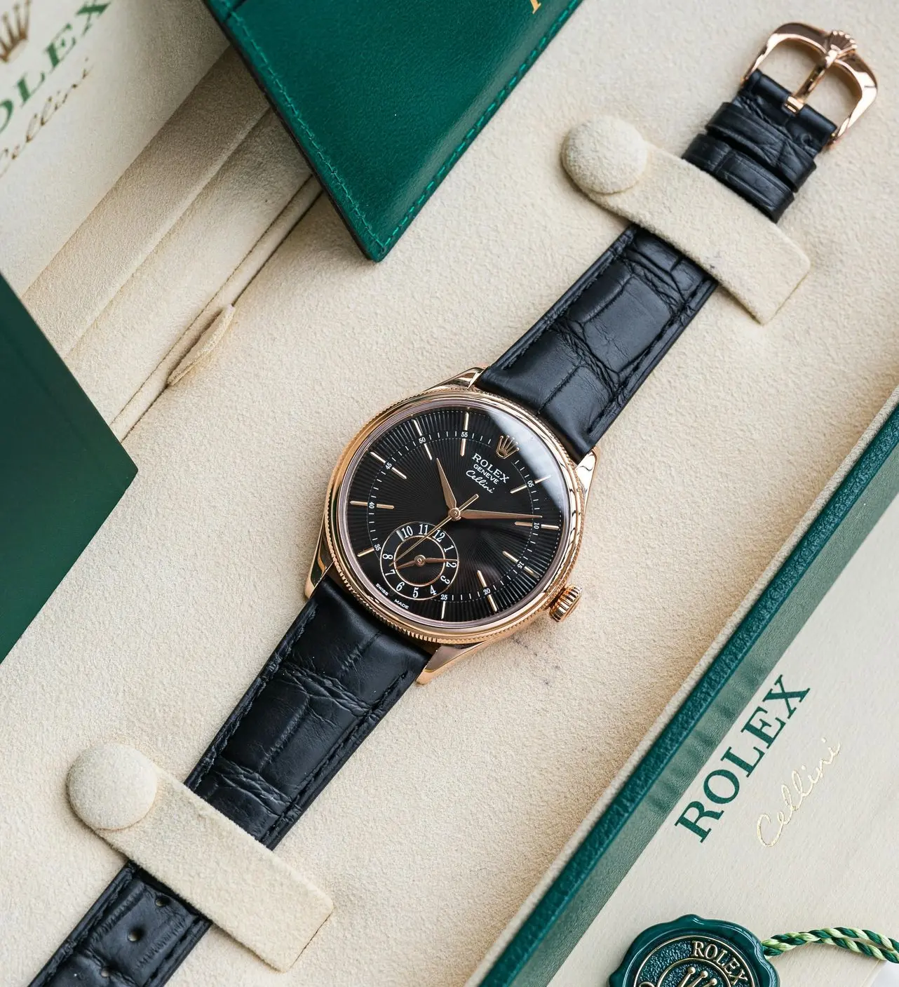 A luxurious Rolex Celestial-style watch with a black dial and rose gold case, paired with a black alligator strap, placed inside a beige presentation box with green Rolex branding.