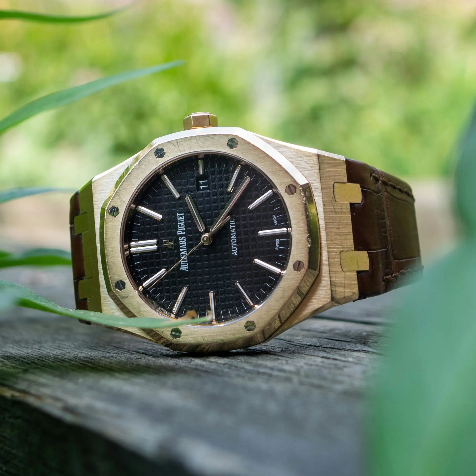 Close-up image of a gold-toned Audemars Piguet Royal Oak watch with a black “tapisserie” dial, brown leather strap, and distinctive octagonal bezel with screws, resting on a wooden surface with greenery in the background.