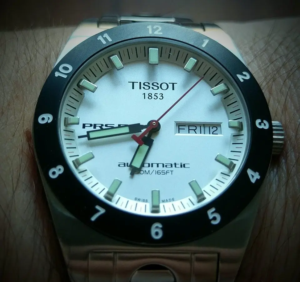Tissot automatic wristwatch with white dial, black bezel, and luminous hands; day-date display; stainless steel bracelet.
