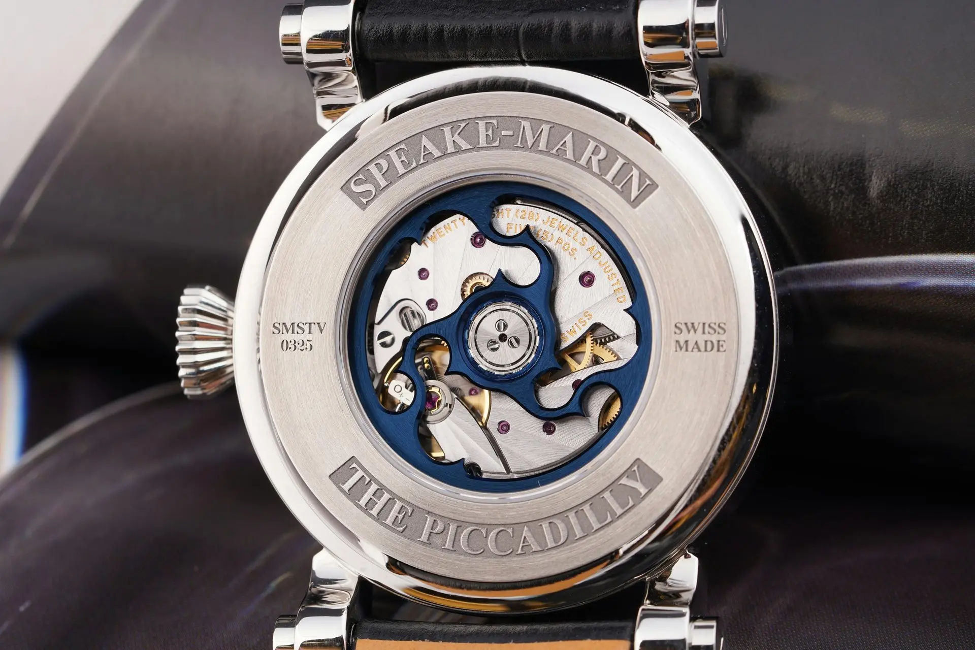 Close-up of a luxury watch back showing an open mechanism with blue accents, engraved branding “THE PICCADILLY” and “SPEAKE-MARIN” on the caseback.