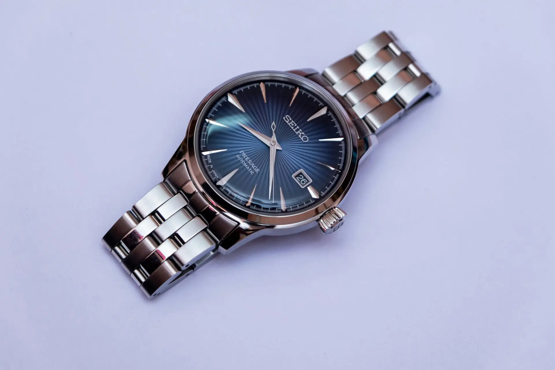 Close-up of a luxury Seiko wristwatch with a blue sunburst dial and stainless steel bracelet, lying on a light purple background.