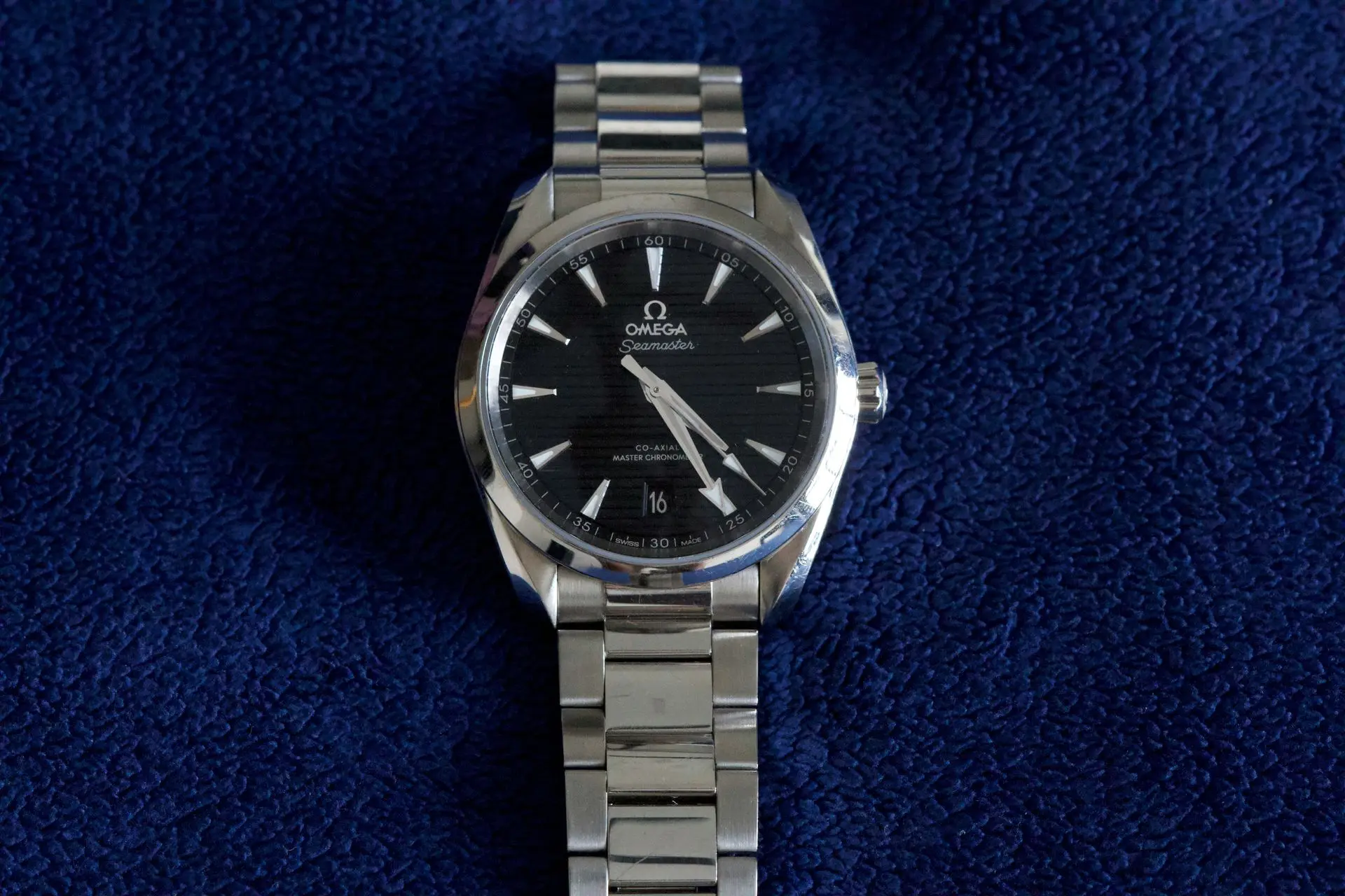 A stainless steel Omega Seamaster wristwatch with a black dial and date display, placed on a textured dark blue surface.