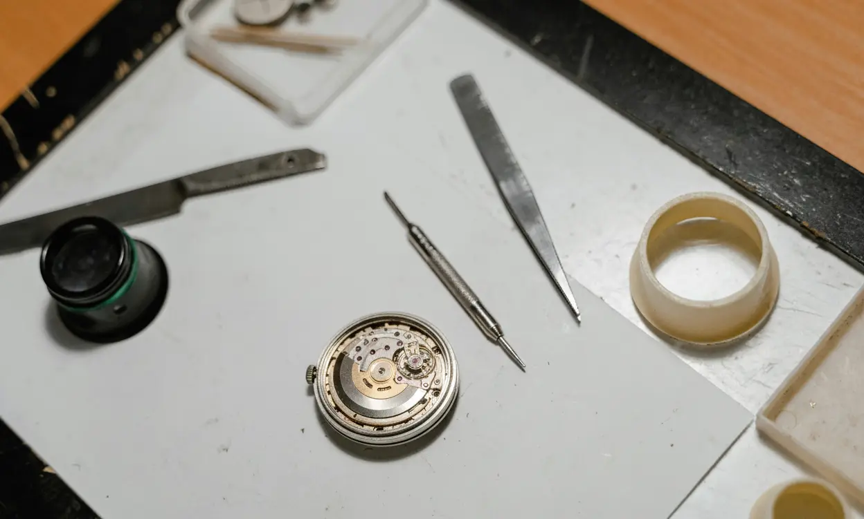 Close-up of an open automatic watch movement on a workbench with precision tools and a roll of tape nearby.