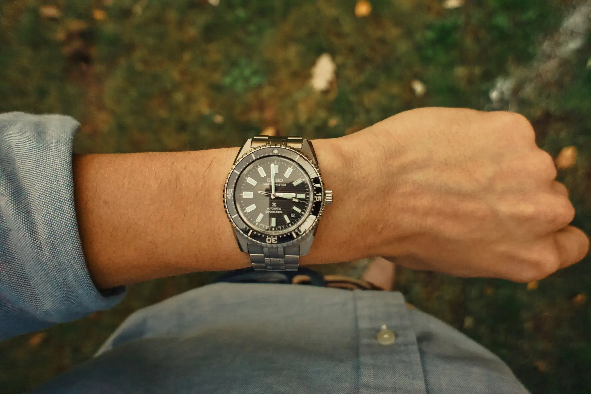 A man's wrist wearing a stainless steel diver's watch with a black dial and rotating bezel, viewed from above against a green outdoor background.