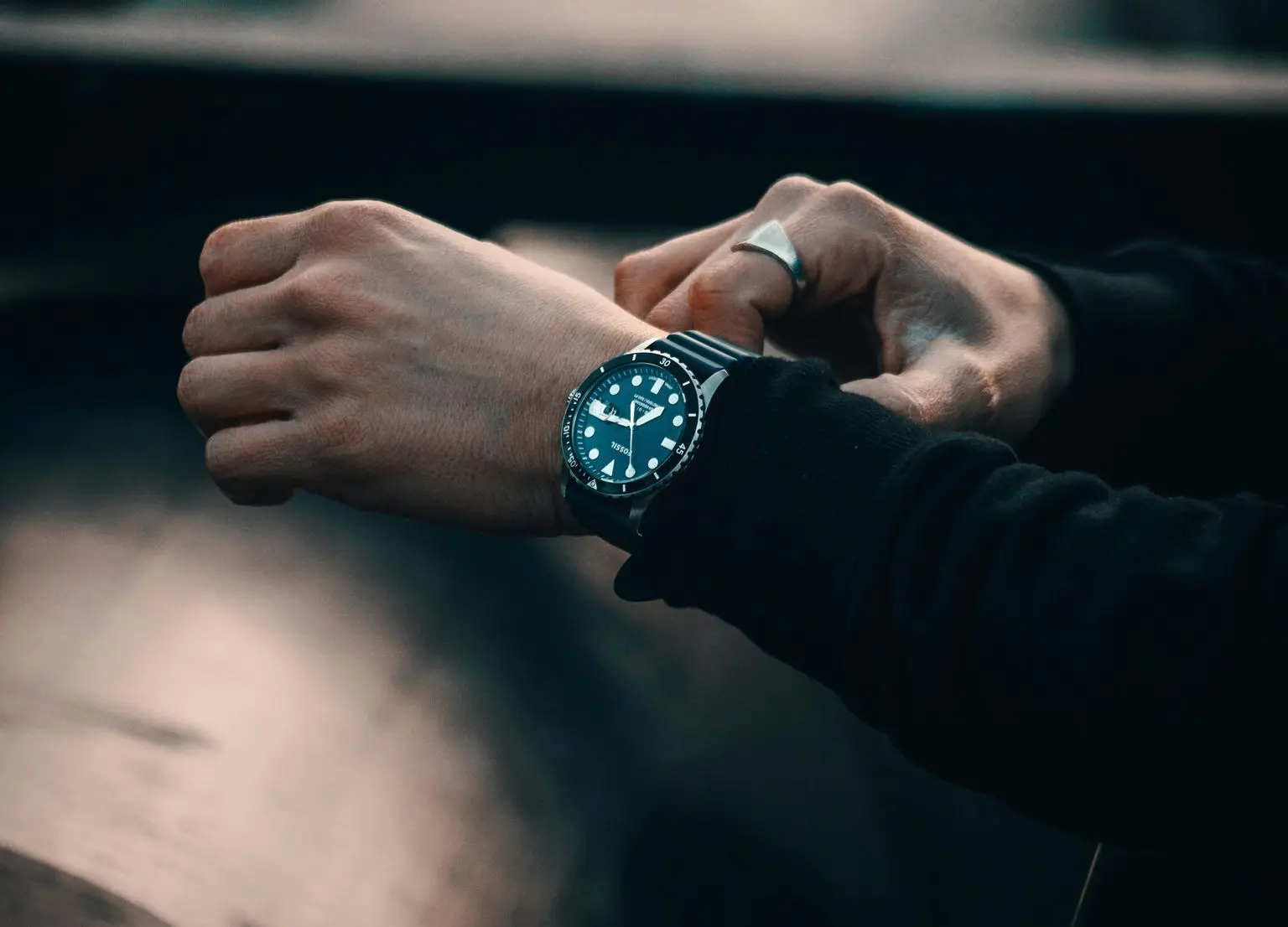 A person wearing a dark long-sleeve top adjusts a stainless steel dive watch with a blue dial and white markers on their wrist.