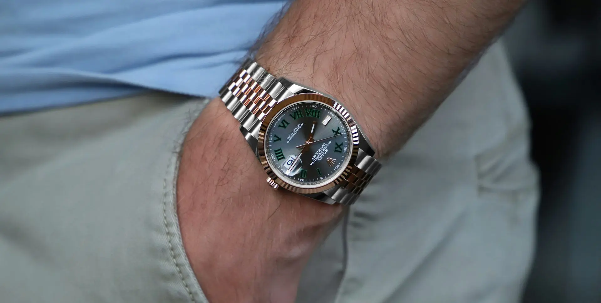  A close-up of a man's wrist wearing a two-tone Rolex-style dive watch with a grey dial, green numerals, and a metal bracelet.
