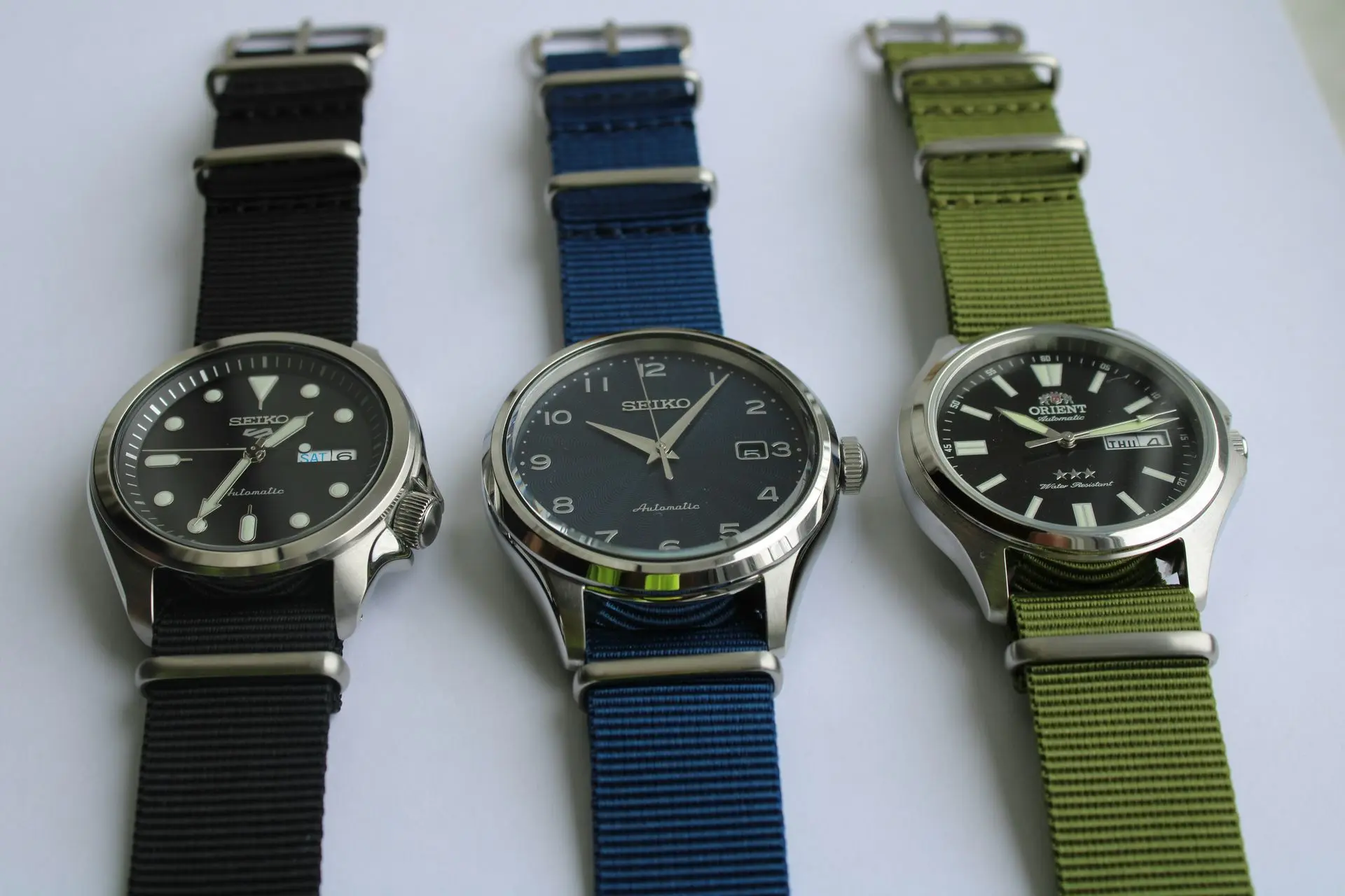 Three watches with nylon NATO watch straps in black, blue, and green displayed side by side