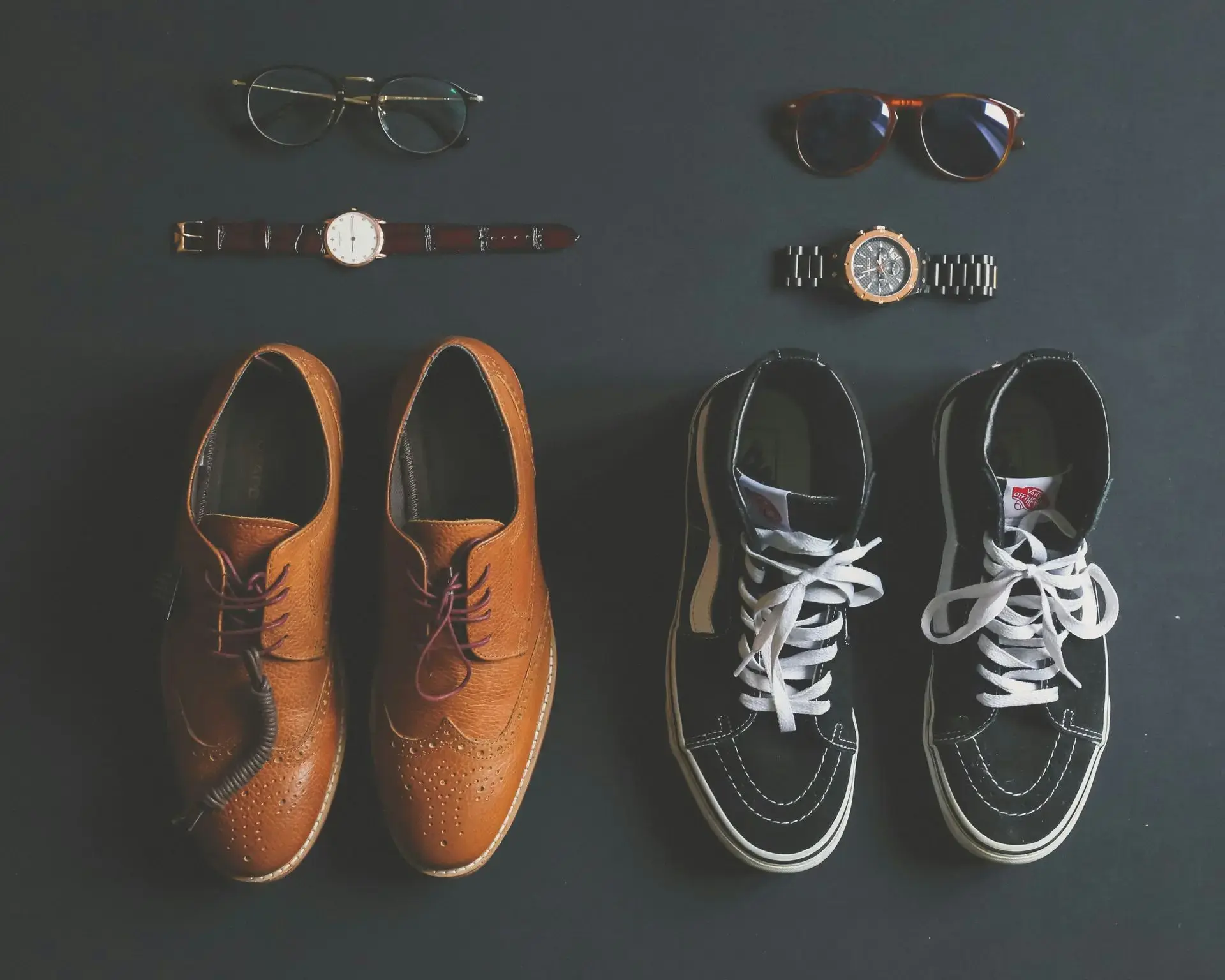 Flat lay of men’s fashion essentials including dress shoes, sneakers, glasses, and two wristwatches illustrating different watch dress codes.