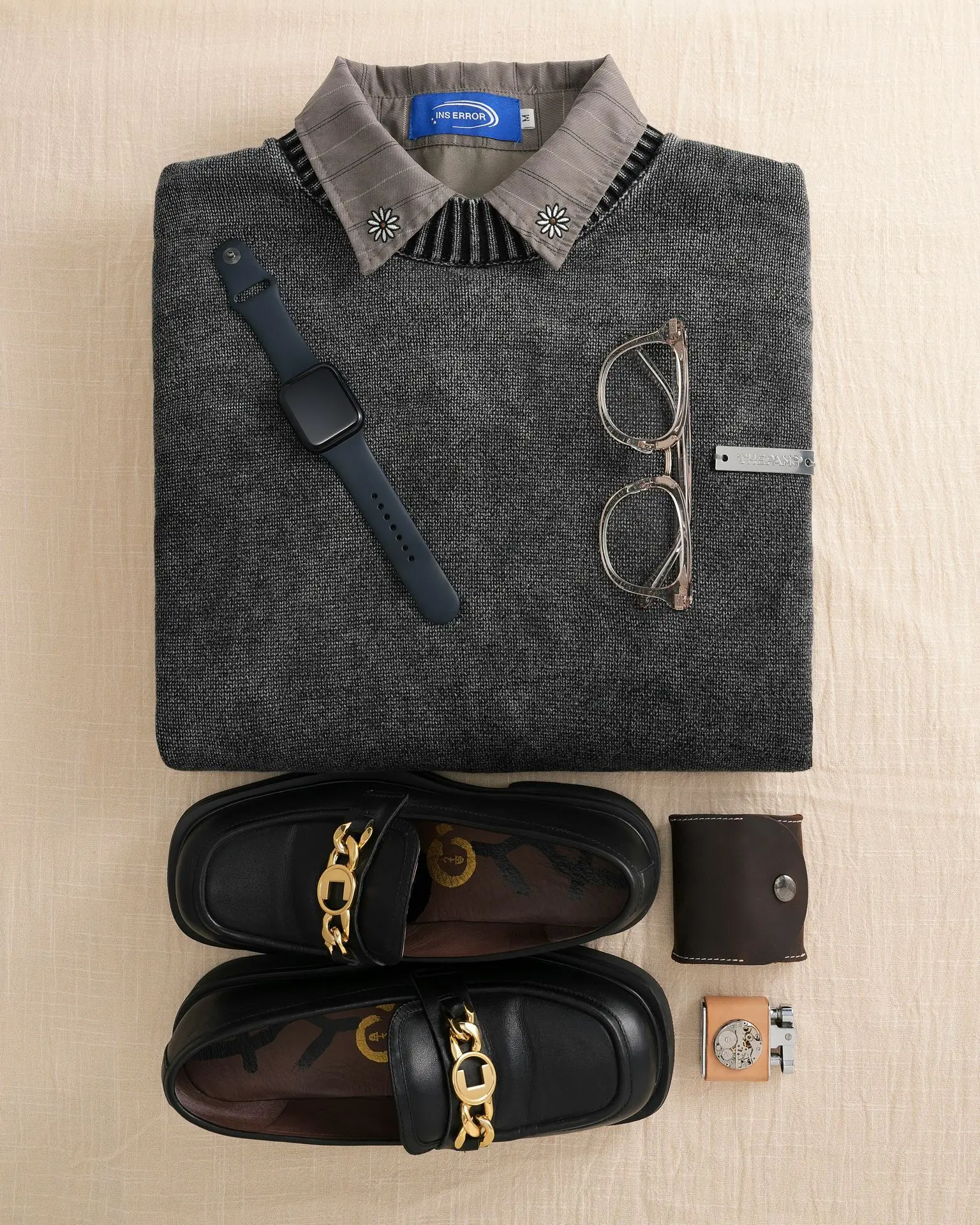 Flat lay of smart casual business essentials including a grey sweater with a collared shirt, black loafers, glasses, a smartwatch, and accessories, illustrating proper watch etiquette for business and smart casual outfits.