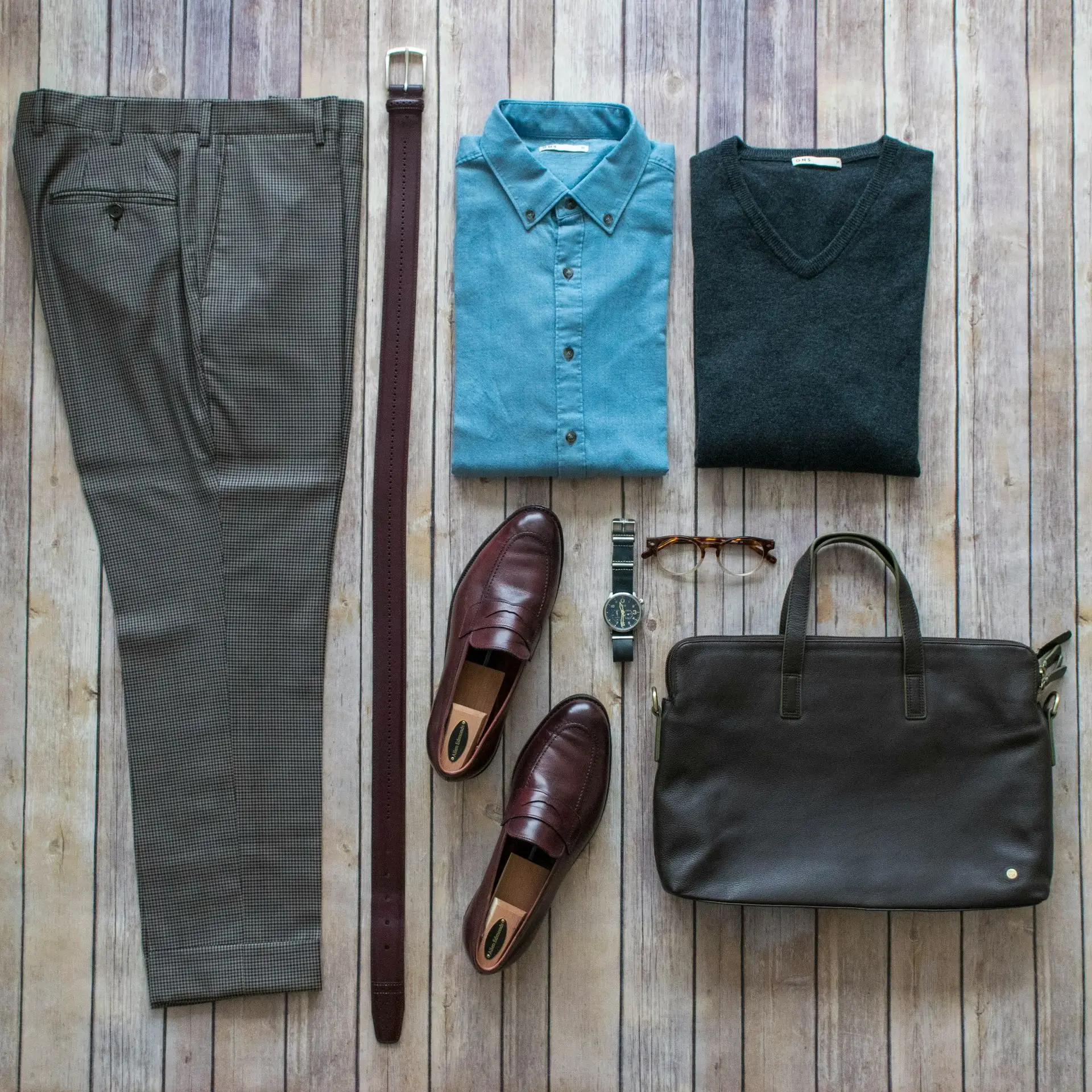 Flat lay of a smart casual outfit including trousers, a denim shirt, sweater, leather shoes, belt, briefcase, and a classic wristwatch, illustrating proper watch etiquette for weddings, job interviews, travel, and other special occasions.