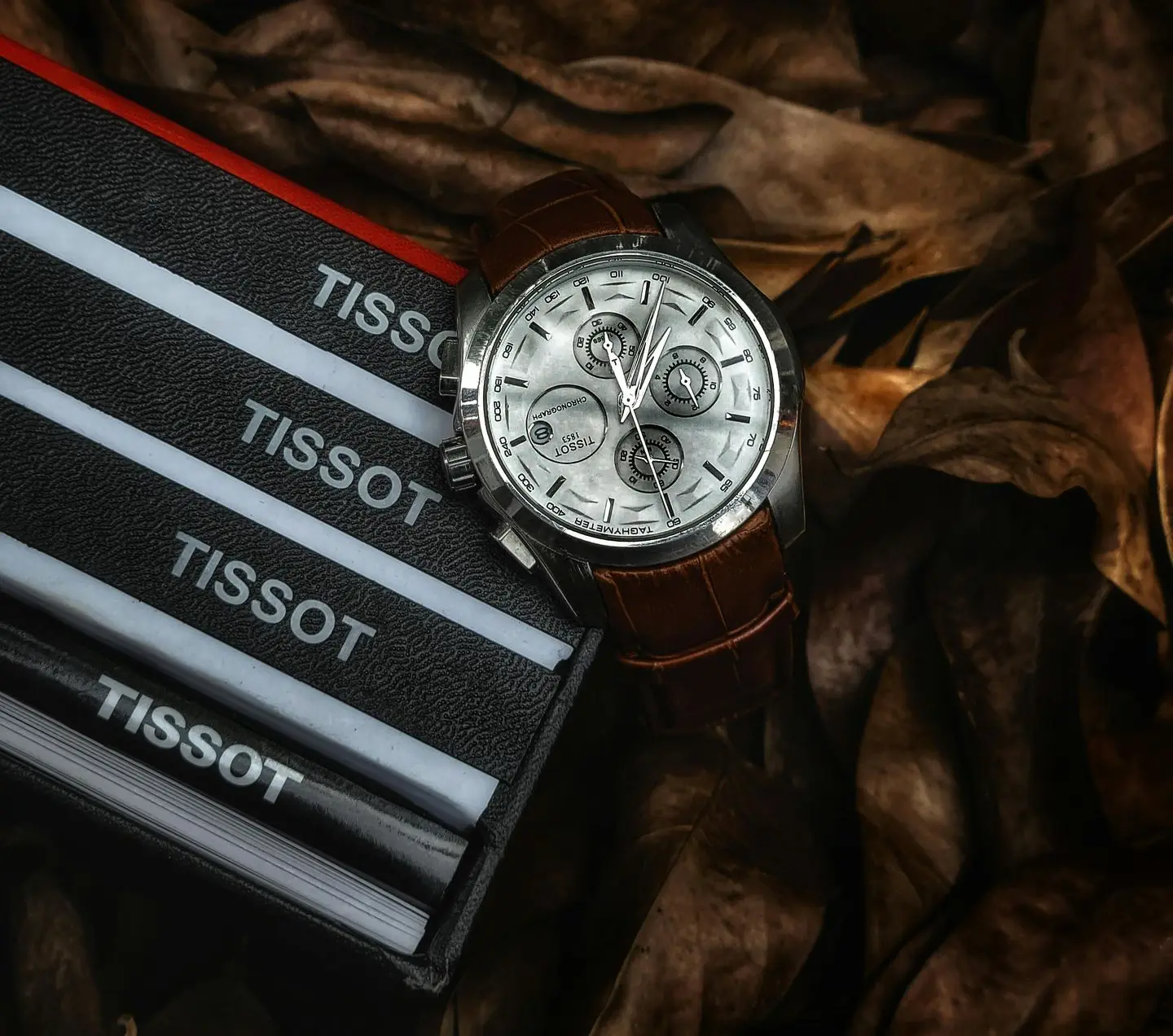 Tissot chronograph wristwatch with brown leather strap placed beside Tissot packaging on dried leaves, symbolizing entry-level luxury purchase.