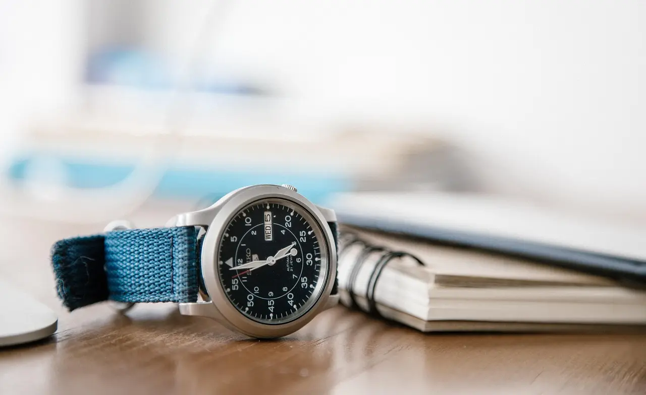 Automatic field watch with blue fabric strap placed on a desk with books in the background.