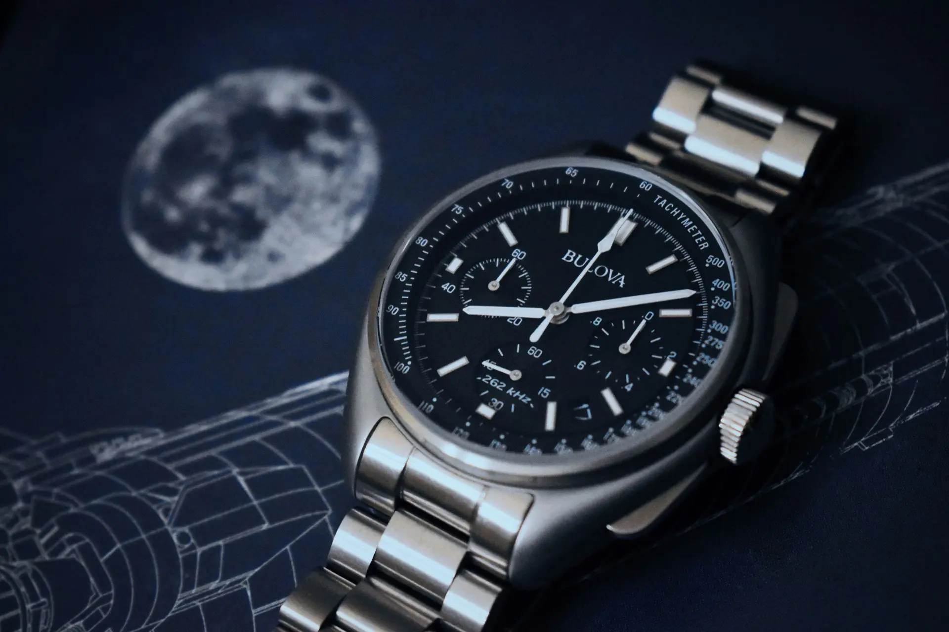 Bulova Precisionist Lunar Pilot chronograph with black dial, tachymeter scale, and stainless steel bracelet against moon background.
