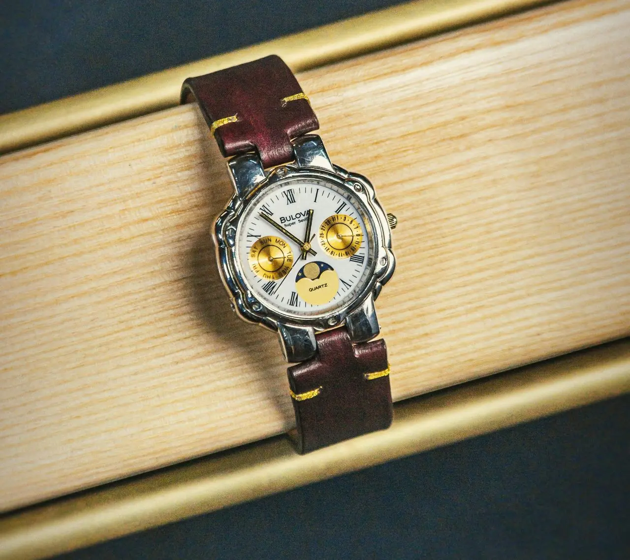 Vintage Bulova quartz moonphase watch with Roman numerals, gold subdials, and brown leather strap resting on wooden surface.