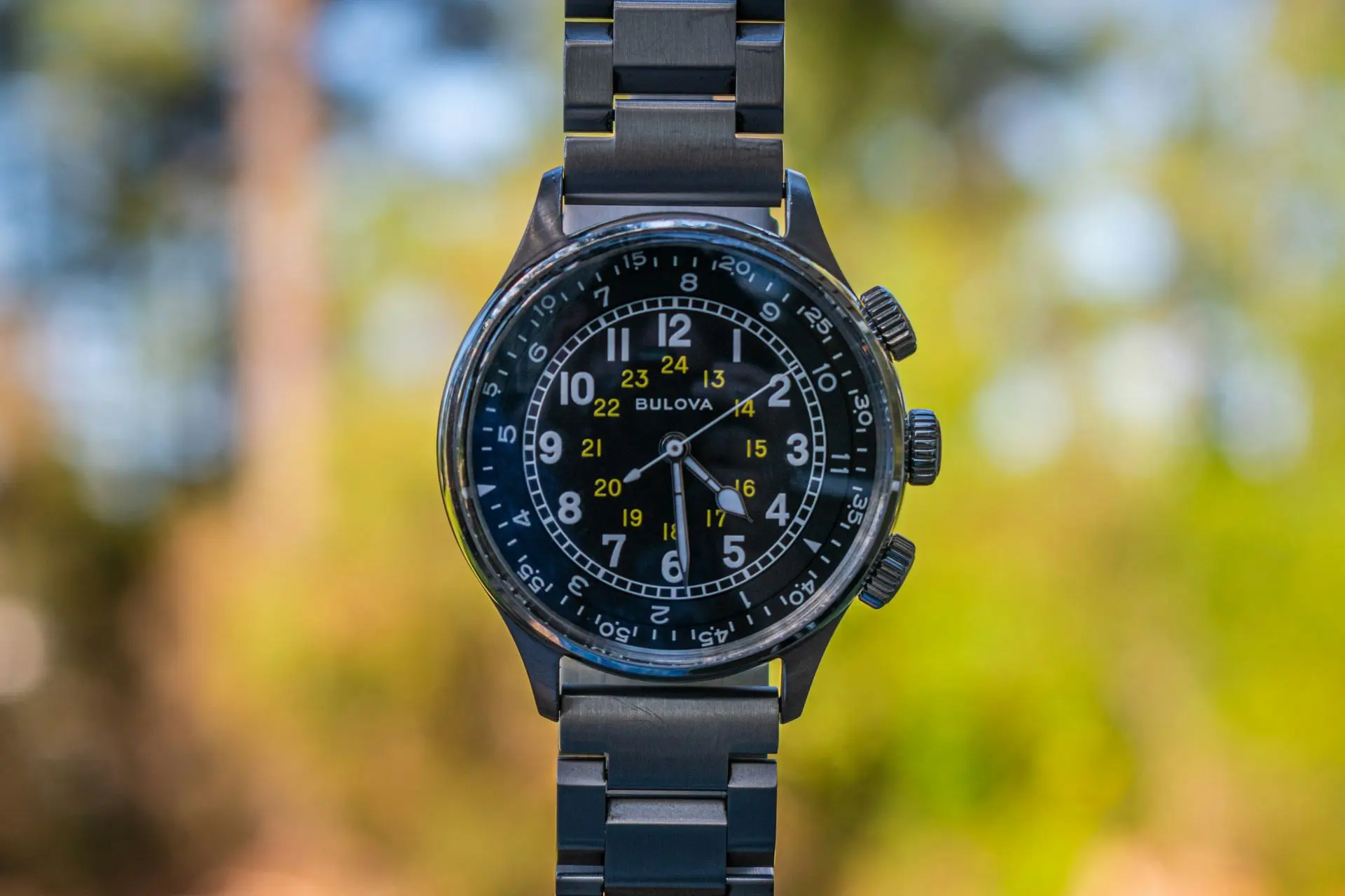 Bulova Hack military field watch with black dial, 24-hour markers, and stainless steel bracelet outdoors.