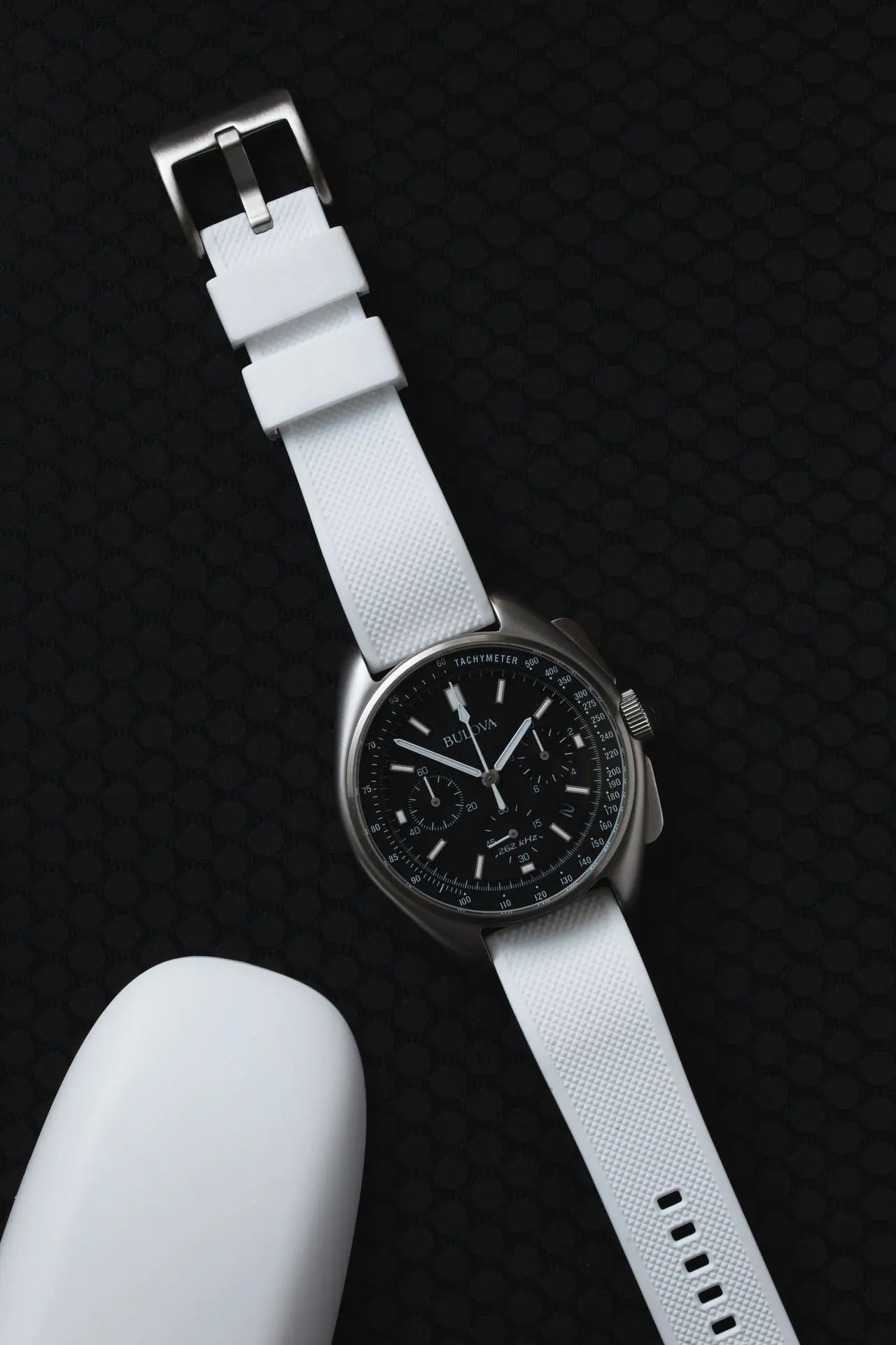 Bulova Precisionist chronograph watch with black dial, tachymeter scale, and white rubber strap on dark background.